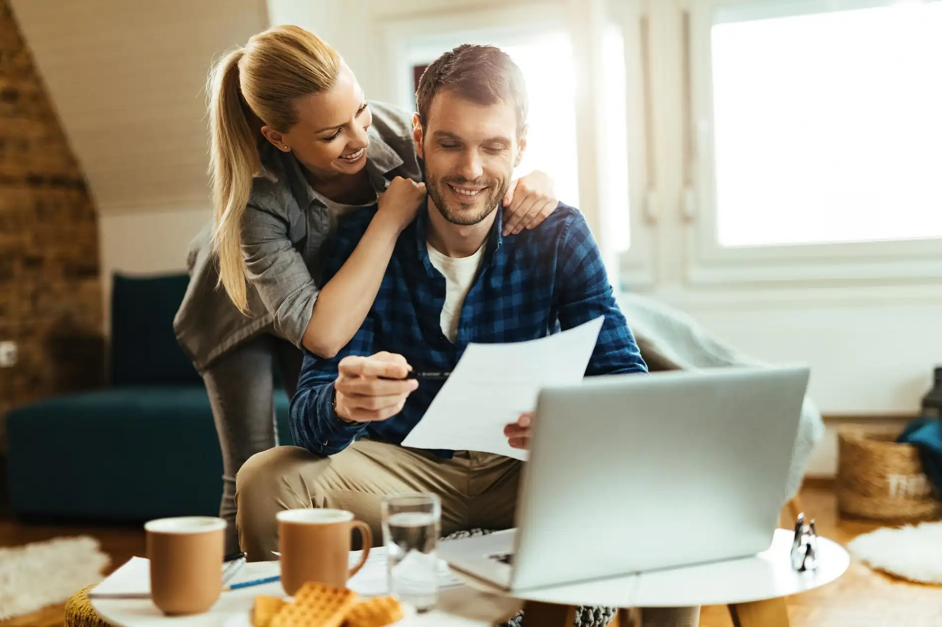 Ein lächelndes Paar sitzt zusammen an einem Tisch, begutachtet Dokumente und arbeitet an einem Laptop, was die gemeinsame Finanzplanung oder die Bearbeitung von wichtigen Unterlagen symbolisiert. Die Frau blickt ihrem Partner über die Schulter, während er ein Formular liest. Sie könnten dabei sein, die Konditionen für ein Erbschaftsdarlehen zu prüfen, um eine anstehende Erbschaft vorzeitig zu nutzen oder sich über die damit verbundenen finanziellen Schritte zu informieren.