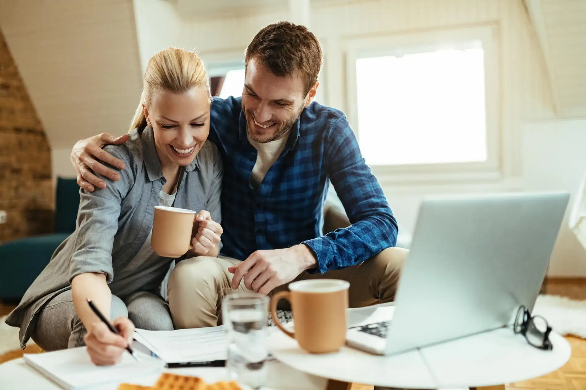 Ein lächelndes Paar sitzt gemütlich zu Hause zusammen und befasst sich mit Unterlagen und einem Laptop, was auf gemeinsame finanzielle Entscheidungen oder die Bearbeitung von wichtigen Dokumenten hindeutet. Die Frau schreibt Notizen, während der Mann sie liebevoll umarmt und mit ihr auf ein Dokument blickt. Diese Szene könnte symbolisch für die gemeinsame Prüfung der Optionen eines Erbschaftsdarlehens stehen, um finanzielle Flexibilität in Bezug auf eine anstehende Erbschaft zu erhalten oder notwendige Formalitäten für die Vorfinanzierung zu klären.