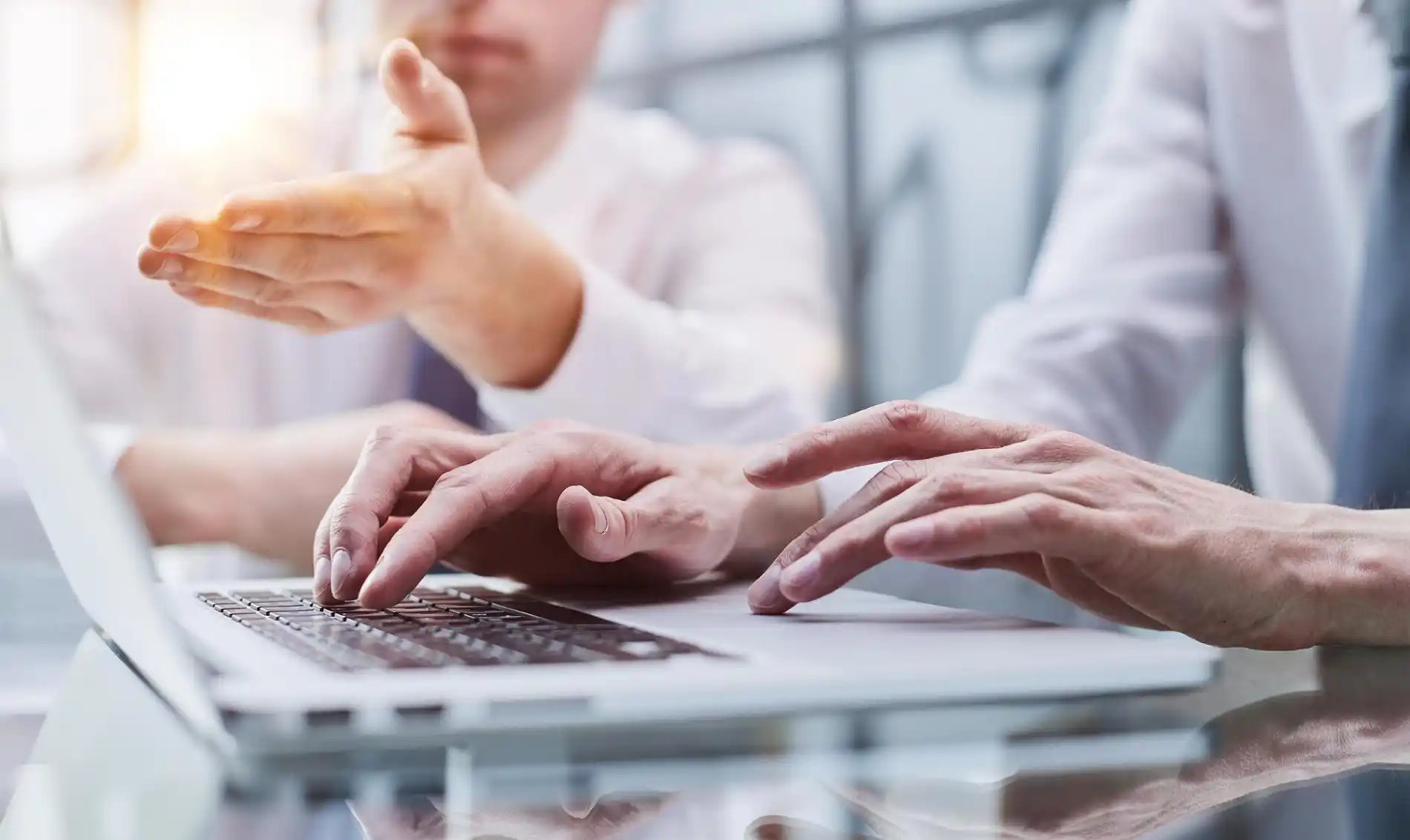 Ein Detailbild zeigt zwei Personen, die an einem hellen Arbeitsplatz zusammenarbeiten. Im Vordergrund tippen die Hände einer Person auf der Tastatur eines Laptops, während die Hand einer zweiten Person auf die Tischfläche zeigt, möglicherweise um einen Punkt auf dem Bildschirm oder einem Dokument zu erklären. Beide tragen weiße Hemden und Krawatten, was auf ein Geschäftstreffen hindeutet. Die Szene symbolisiert die Analyse von Geschäftszahlen und Finanzergebnissen, die direkt mit der Führung eines Ertragskontos verbunden ist.