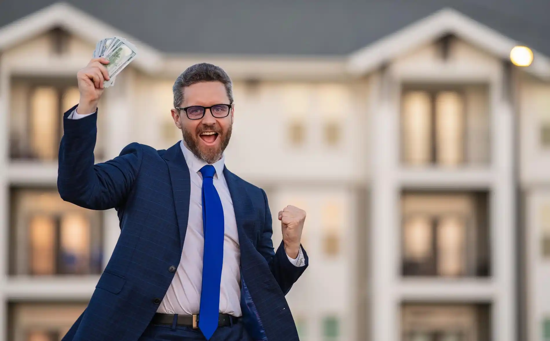 Ein überglücklicher Mann in einem Anzug mit Brille und blauer Krawatte jubelt vor einem großen, modernen Mehrfamilienhaus. Er reckt die Faust in die Luft und hält triumphierend einen Fächer von Geldscheinen in der anderen Hand. Das Bild vermittelt ein Gefühl von Erfolg und Wohlstand. Das Mehrfamilienhaus im Hintergrund symbolisiert die Investition in Realkapital, während das Geld für den Gewinn aus dieser Investition steht.