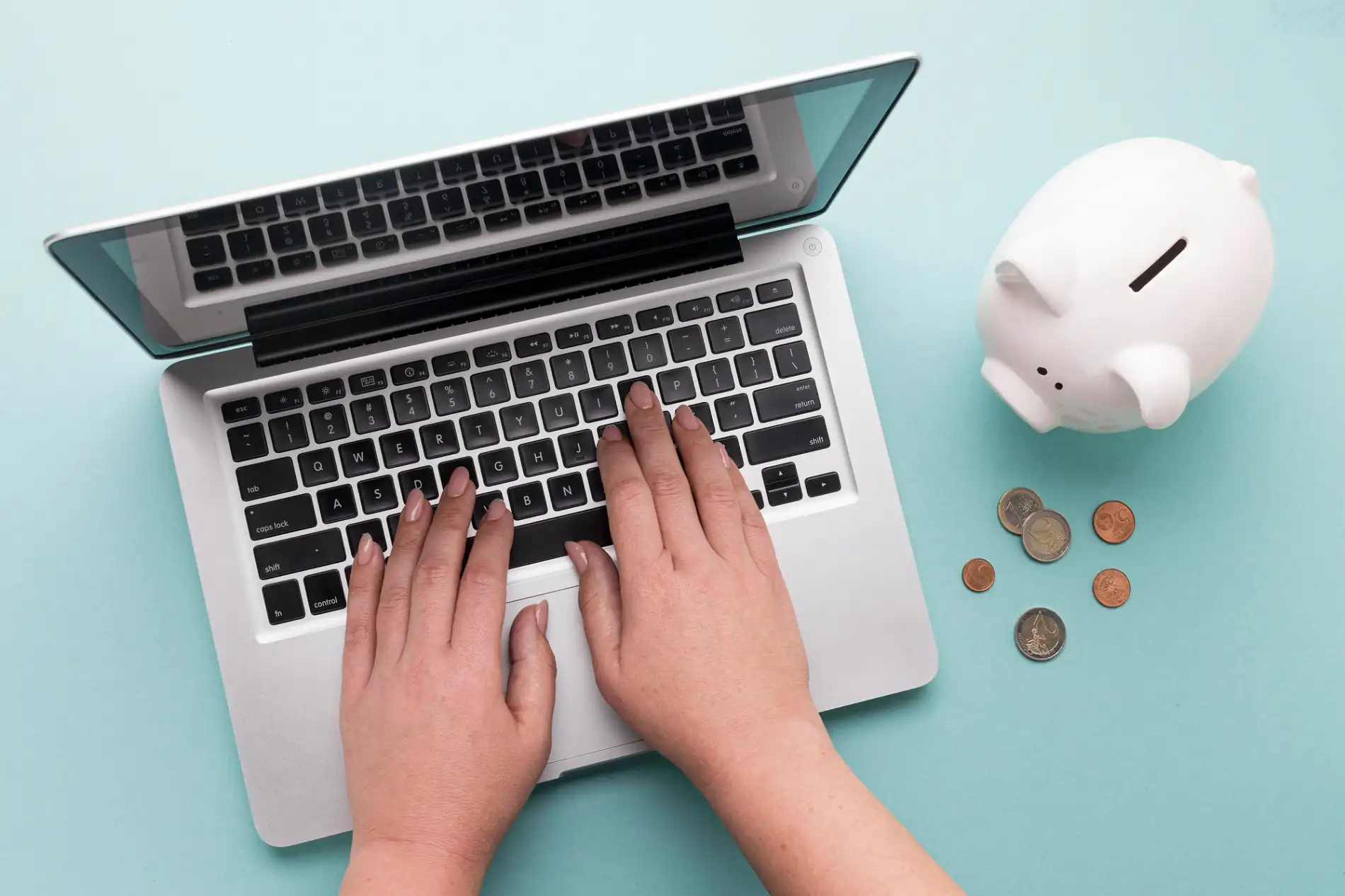 Ein Flat-Lay-Bild zeigt eine Person, deren Hände auf einer Laptop-Tastatur ruhen. Rechts neben dem Laptop steht ein weißes Sparschwein, aus dem einige Münzen gefallen sind und auf der türkisfarbenen Oberfläche liegen. Das Bild symbolisiert die digitale Verwaltung von Finanzen und veranschaulicht visuell den Unterschied zwischen Kapital und Vermögen: Das Sparschwein repräsentiert das passive Vermögen, während die aktive Nutzung des Laptops für Finanzgeschäfte die Umwandlung von Vermögen in produktives Kapital darstellt.
